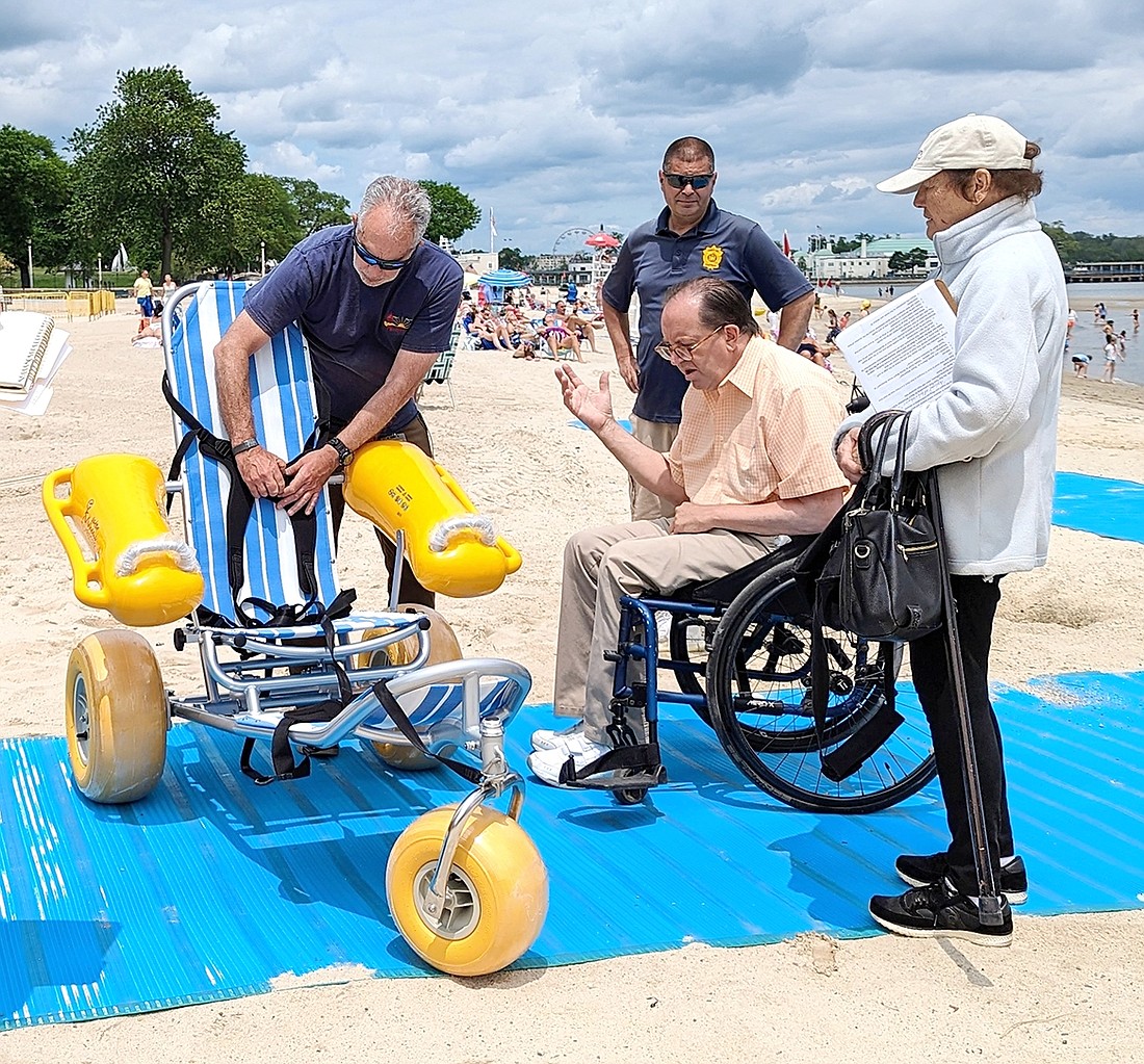 Standing on a new ACCESSMAT that heightens wheelchair accessibility on Oakland Beach at Rye Town Park, Park Director Russell Gold (left) unfastens a belt so ADA specialist and advocate Michael Hellmann can try out one of the new beach wheelchairs offered at the property. Park Ranger Captain Michael Cancel and Judy Hellmann stand by.