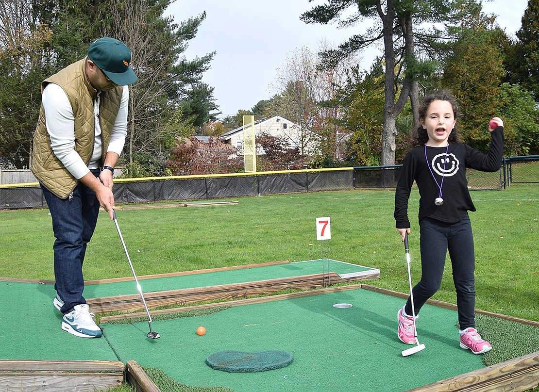 Ridge Street School kindergartener Blake Laskin triumphantly holds up her ball after sinking the seventh hole. Her dad, Michael, lines up to take another swing.