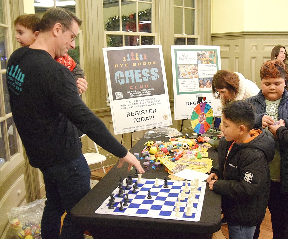 Professional Chess Instructor Brian Wolff (left), with the Rye Brook Chess Club, holds his 2-year-old son Mason while playing a friendly game with Port Chester resident Dave Javier, 8.