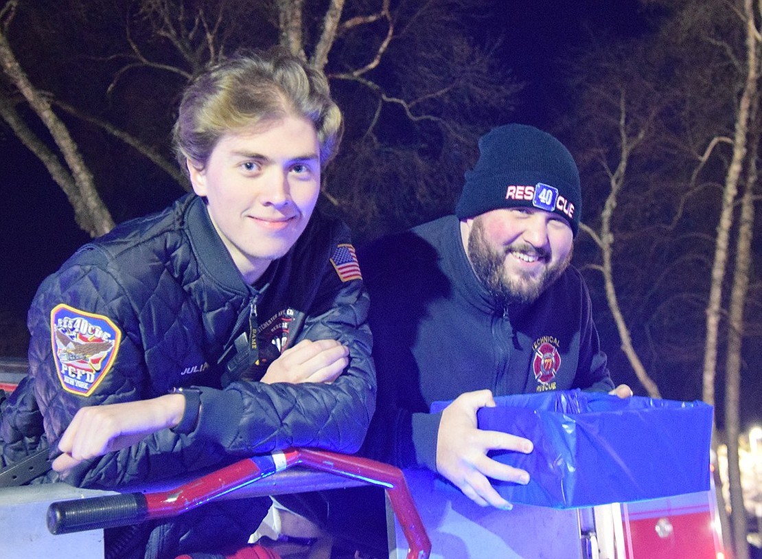 Port Chester firefighters Julian Silva (left) and Matt Ganz smile for the camera in the bucket of a firetruck. Ganz holds a box full of Chanukkah gelt, preparing to drop it for the children.