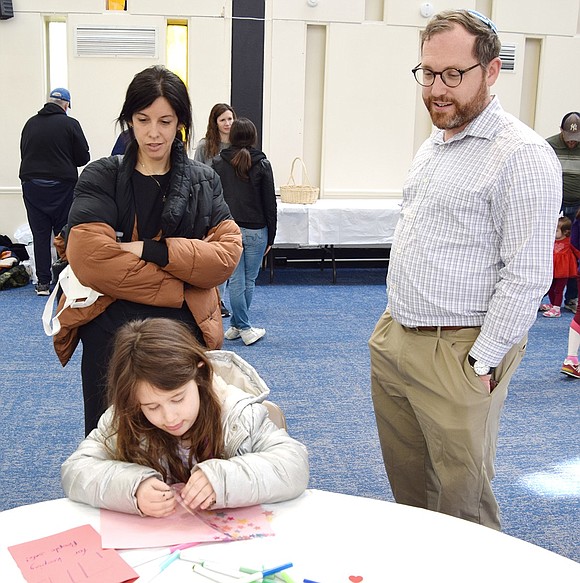 Five-year-old Talia Balsam, a kindergartener at Ridge Street School, writes a letter to an Israel Defense Forces soldier while her mother Sarah and Rabbi Ben Goldberg take a peek at her work.