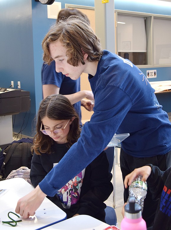 Blind Brook High School freshman Sasha Harra (standing) distributes batteries, wires and pipe cleaners to 11-year-old Arya Elliott for her to construct her BristleBot.