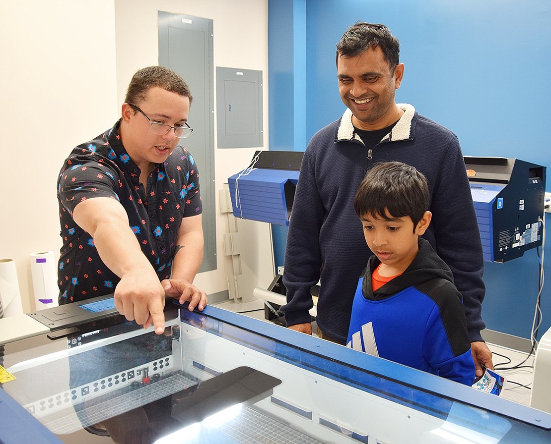 Fab Lab Manager Josh Colon (left) explains how students use the laser engraver for their work to Millenium Place resident Praveen Prathy and his son Rohan, a first-grader at Ridge Street Elementary School.