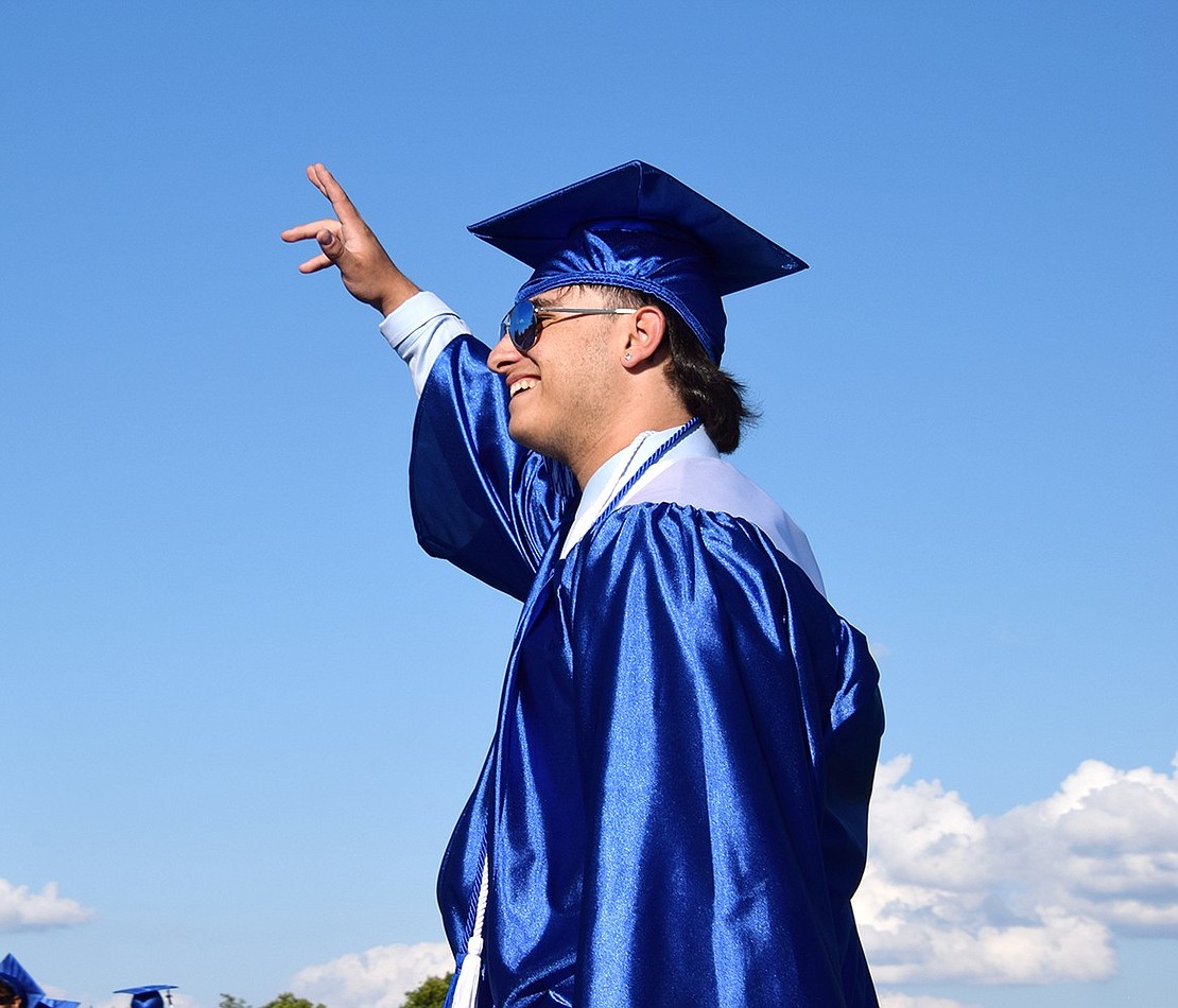 John Tomassetti’s dark blue gown contrasts with the aqua skies as he waves to loved ones in the bleachers.