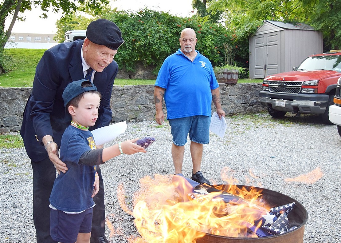 Chester “Chet” Edwards, a veteran and member of the Knights of Columbus who served as emcee for the ceremony at 327 Westchester Ave., guides 5-year-old Rye Brook resident Maxwell Fischer in disposing of a flag as Grand Knight Mark Scocchera watches.