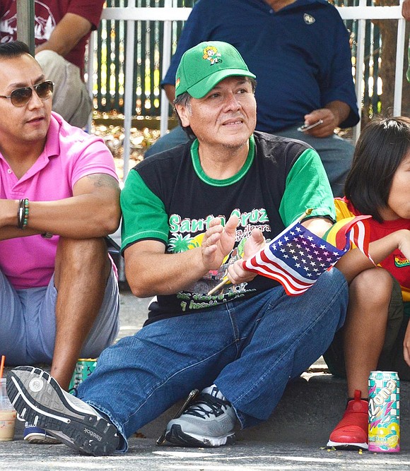 It’s hard to wave the American and Bolivian flags while clapping, but Osvaldo Castillo of Poningo Street, watching the parade from in front of Irv’s Mini Mart, does his best. 