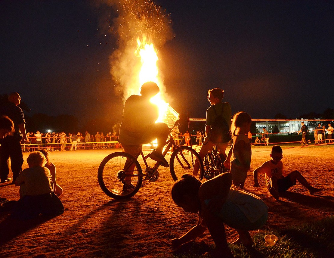 Captivated audience members are silhouetted by the warm orange glow of the bonfire. 