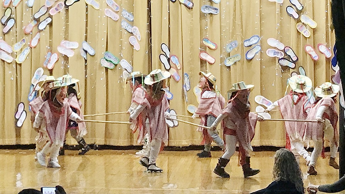 Adorned in Mexican masks and outfits, kindergarteners perform a circular dance on a brightly lit stage.  Courtesy of Carolee Brakewood 