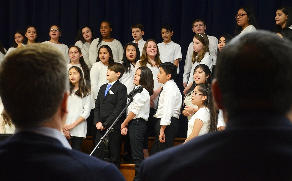 As seen between the heads and shoulders of Port Chester Trustees Dan Brakewood and Luis Marino, the Port Chester Middle School 7th Grade Chorus leads the audience in singing “We Shall Overcome” with a variety of soloists.