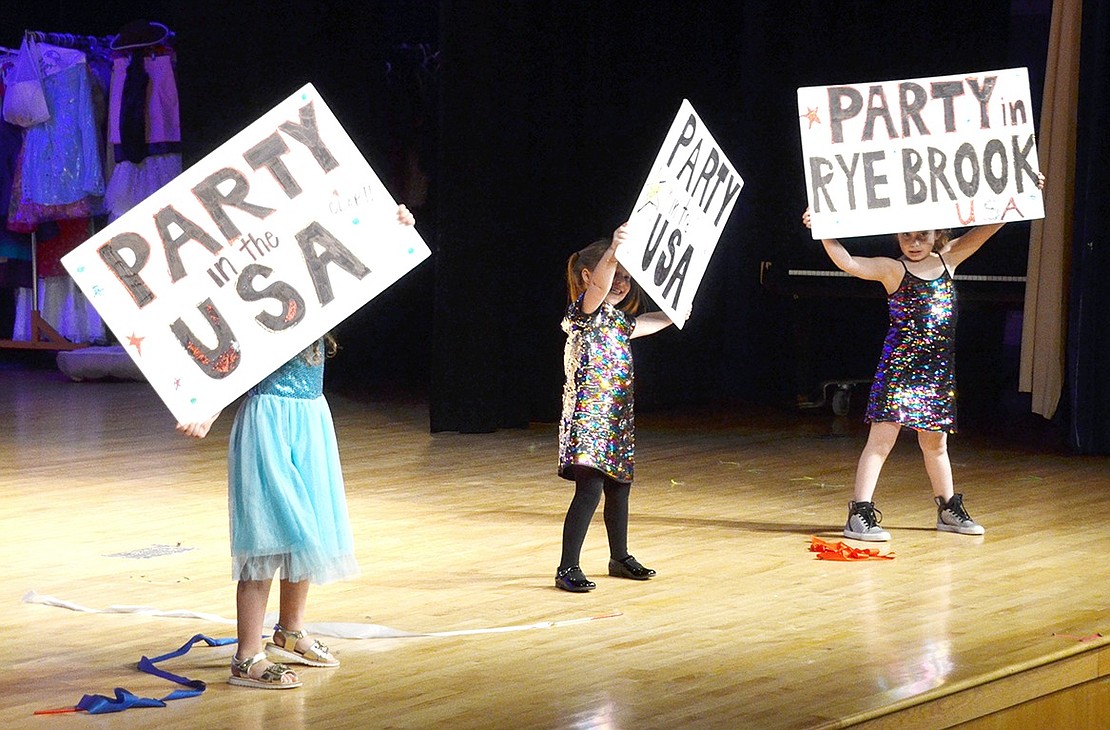 The Girly Girls, kindergarteners Clara Mensch (left), Sammy Capon and Zoey Cohn, hold up signs during their dance to Miley Cyrus’s “Party in the U.S.A.” to let the auditorium know the real party is in Rye Brook.