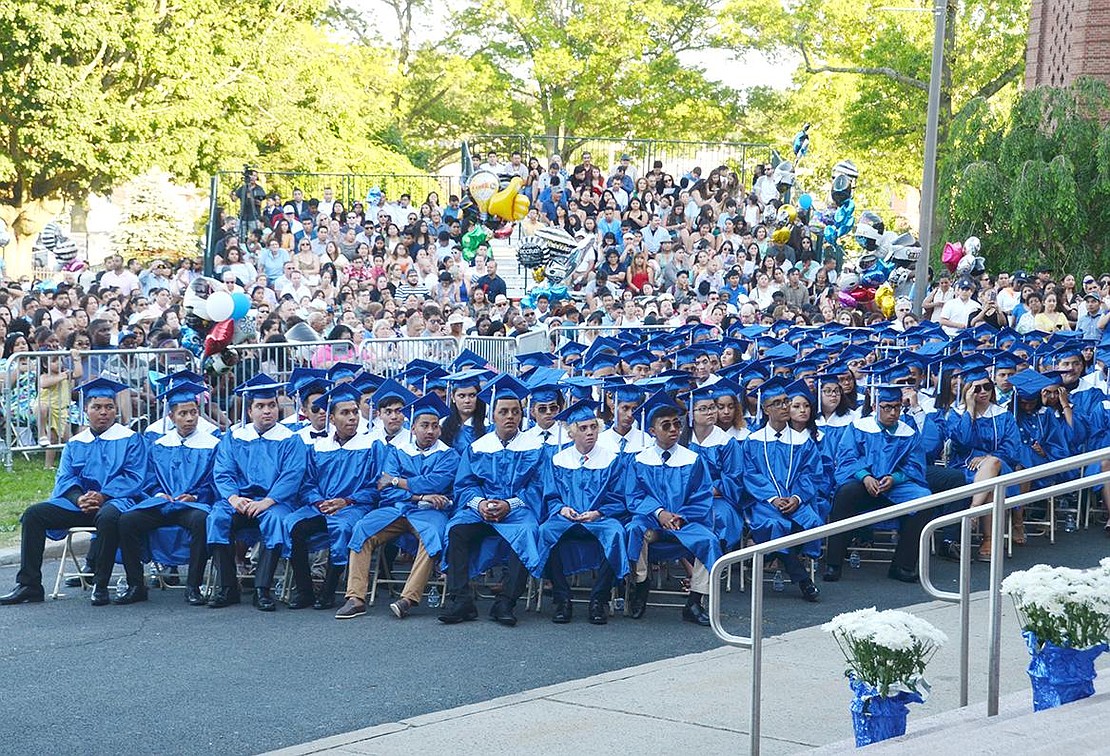 Half of the Class of 2019 is shadowed by friends and family in bleachers and the barricaded area behind them as they sit in the flag plaza for the first graduation ceremony off the football field.