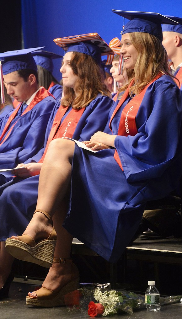 Julia Short flashes a charming smile as she sits with her classmates, all adorned in caps and gowns, on the stage.