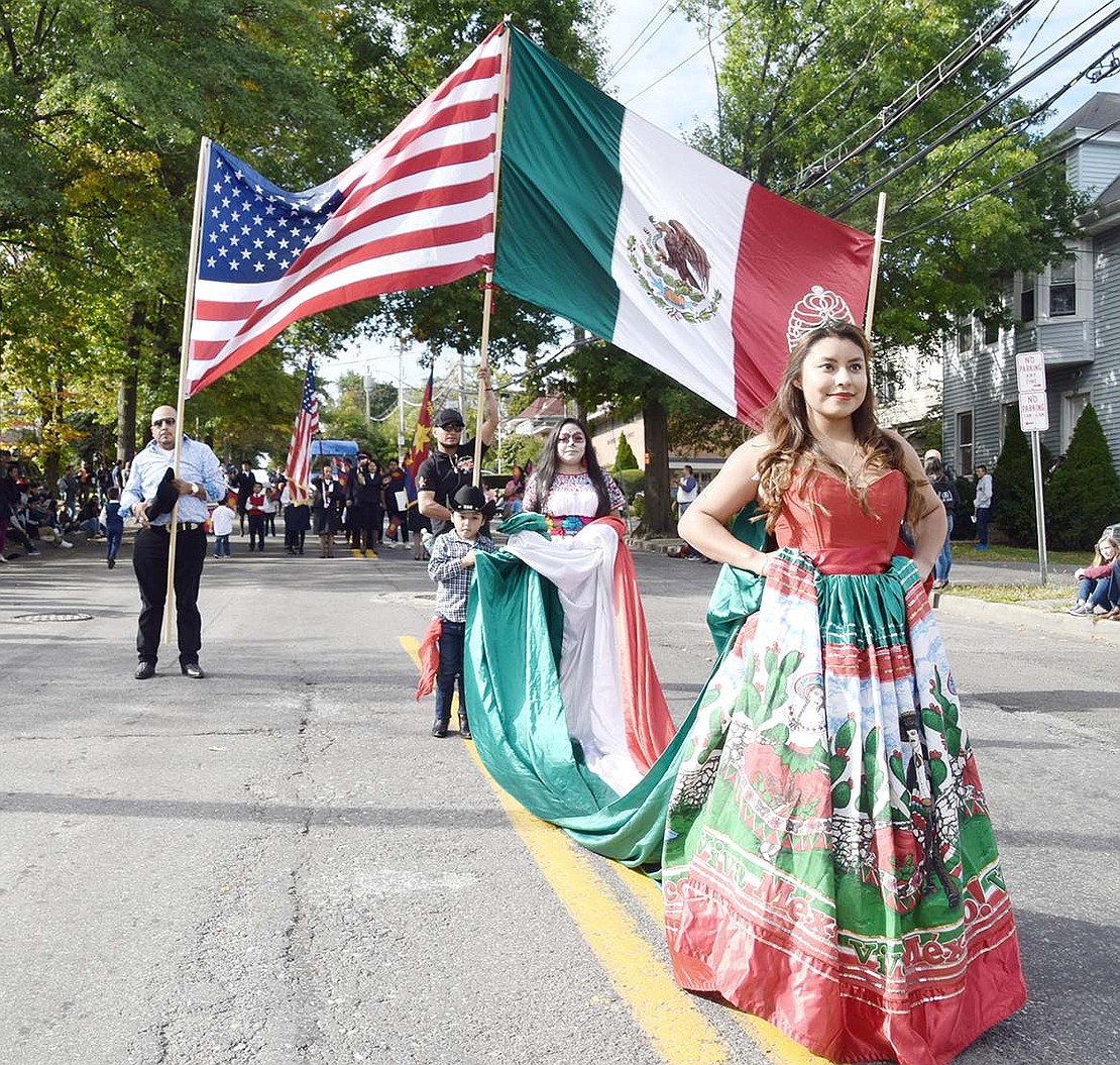 With pretty tiara and long Mexican dress, Oak Street resident Leslie Cruz, 23, walks in front of two large conjoined Mexican and American flags during the procession.