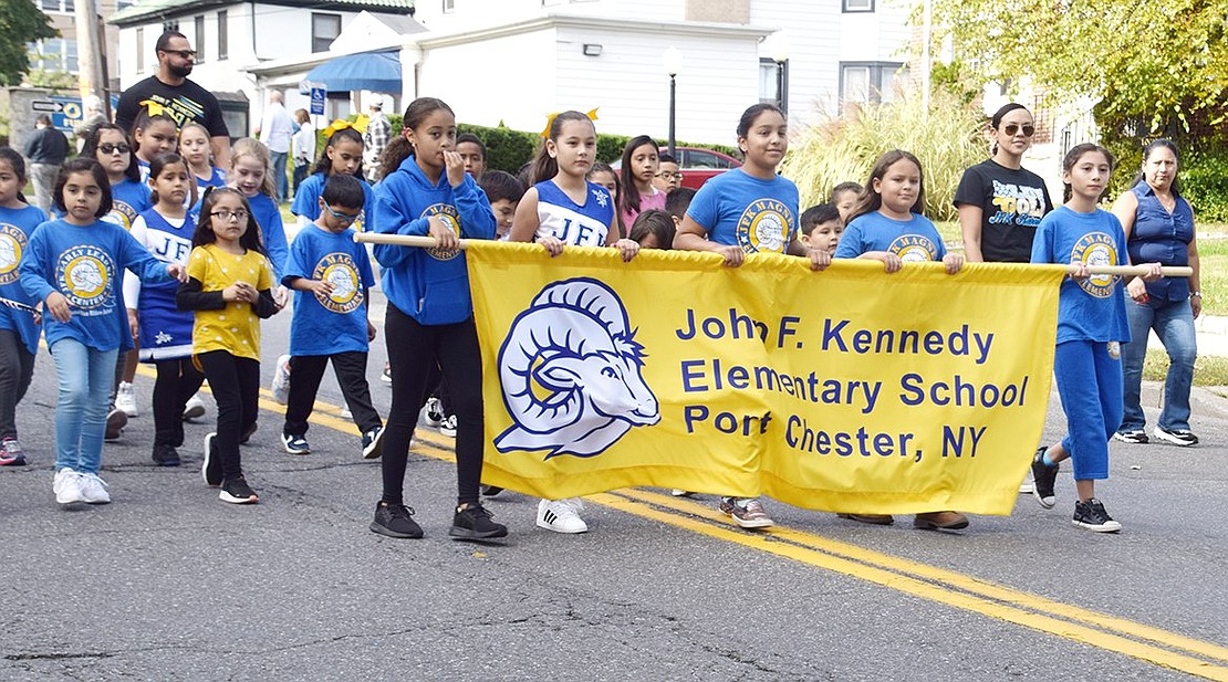 Dozens of students of all ages march down the street together while representing John F. Kennedy Elementary School.