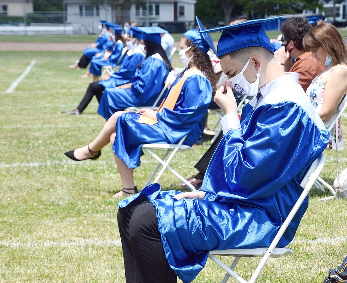 While graduates and parents take a moment of silence for the lives lost to COVID-19 and violence, Luis Gallardo bows his head and tightens his commemorative face mask.