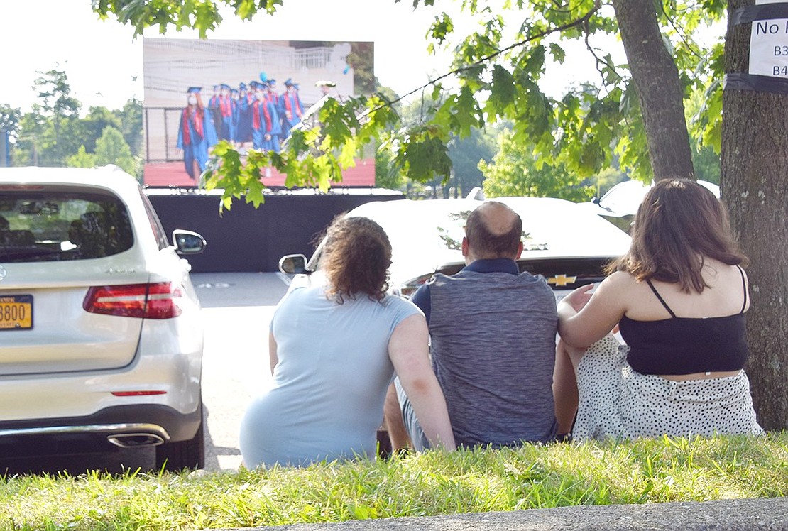 Like for a drive-in movie, Adena (left), Mark and Aviella Kibel find a shady spot on a hill between parking spaces to watch the graduation ceremony live streamed on a big screen. While families were not allowed in the stadium, they could still park nearby and watch.