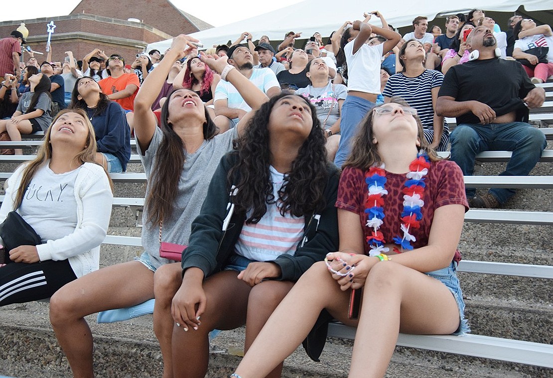From left, Mary Ann Castro, Michelle Rabang, Maya Morales, 13, and Catiana Dias, 12, all of Greenwich, as well as all the people in the stands behind them, look up to watch as skydiver Jeff Provenzano descends from the sky above Port Chester High School’s Ryan Stadium. His performance was a novel highlight of the local Fourth of July festivities this year.