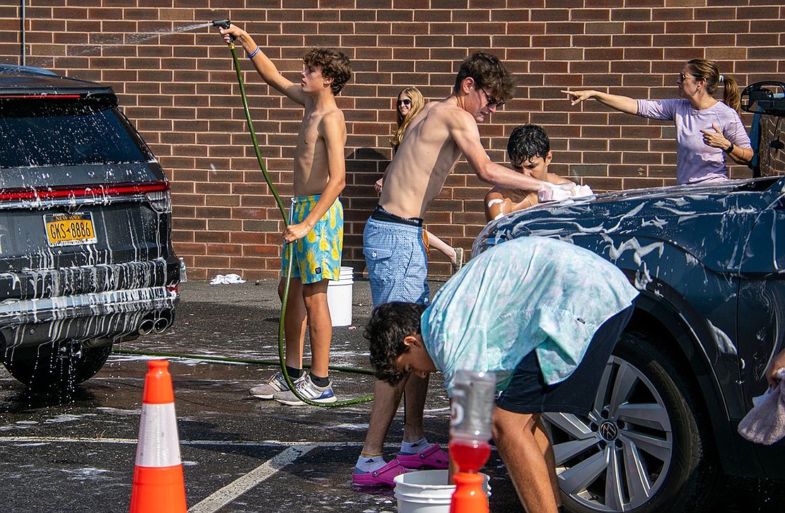 Junior midfielder Eli Zimmerman washes the hood of an SUV with a sponge, while sophomore goalie Luka Cuk bends down to replenish the water in his own sponge. Behind them, Dylan McRedmond, a sophomore midfielder, hoses down another SUV, and moms direct their boys as they work the line.