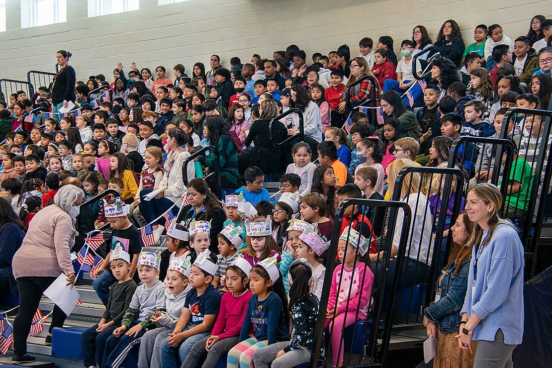Students and teachers sit in the bleachers in the gym of King Street Elementary School, decorative Veterans Day-themed paper crowns adorning some of their heads while others wave American flags.