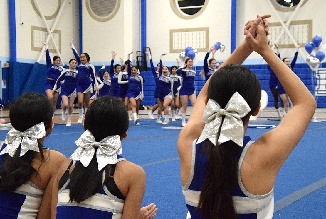 Sitting on the sidelines, varsity squad members root on the junior varsity team as they rambunctiously jog to their positions on the mat to perform their game day routine.